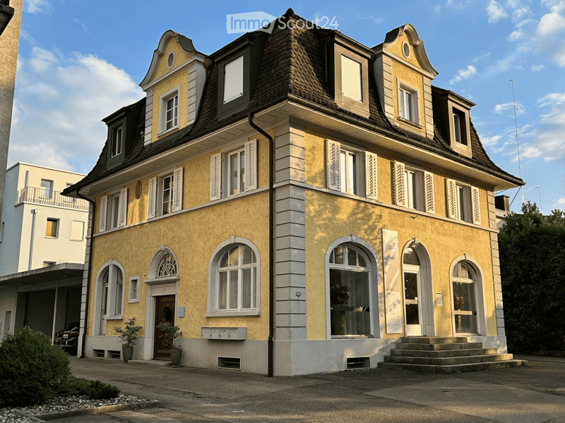 3-story yellow building, arched windows, black roof, white trim, steps at entrance, small balcony on 2nd floor, car parked nearby
