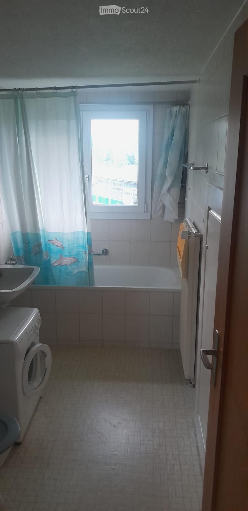 Bathroom with a bathtub, sink, shower curtain, radiator, and washing machine. The room has tiled walls and floor, and a window above the bathtub.