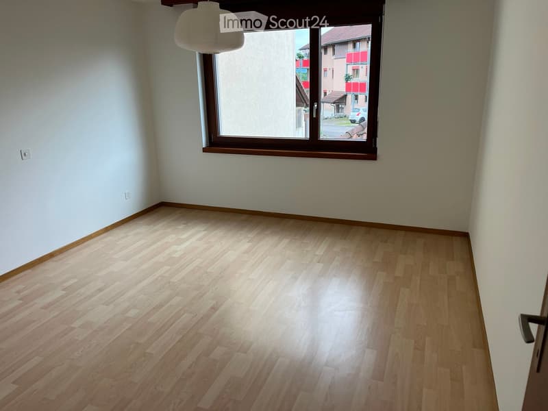 Empty room with light wooden flooring, white walls, and a large window with wooden frame. Natural light illuminates the space.