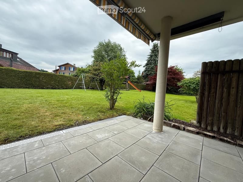 Patio area with tiled flooring, covered by a striped awning, overlooking a well-maintained lawn with trees, a swing set, and a small playhouse in the background.