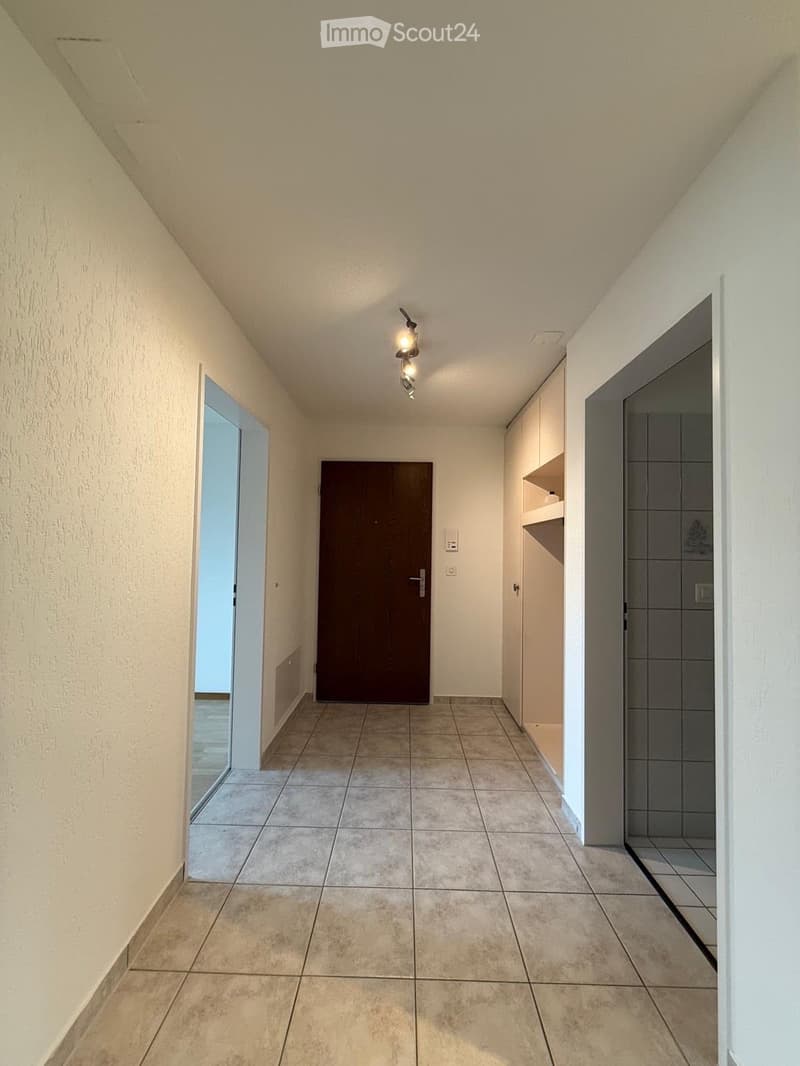 Empty hallway with tiled floor, white walls, and a single light fixture on the ceiling. Features a closed wooden door and an open doorway leading to another room.