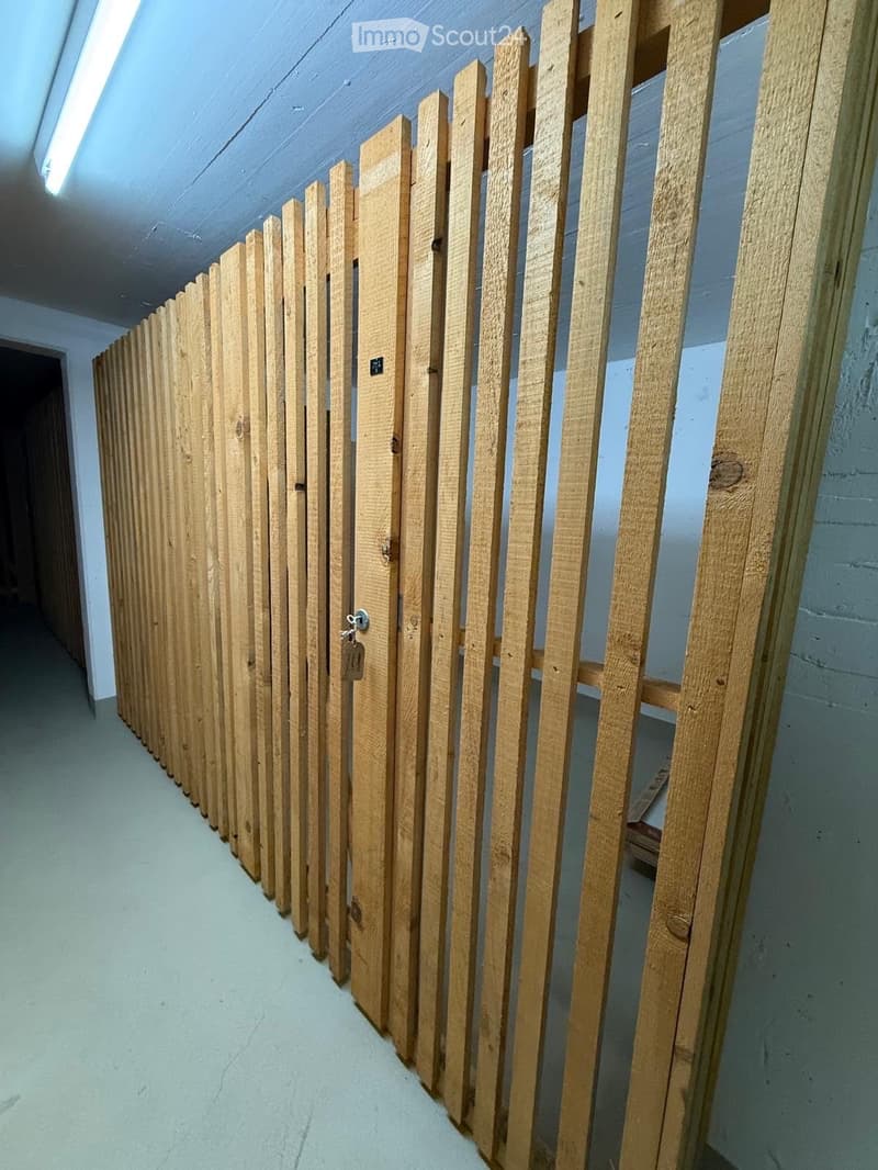 A long, narrow hallway with a wooden fence-like structure on one side, featuring vertical wooden slats and a small door with a lock and tag.