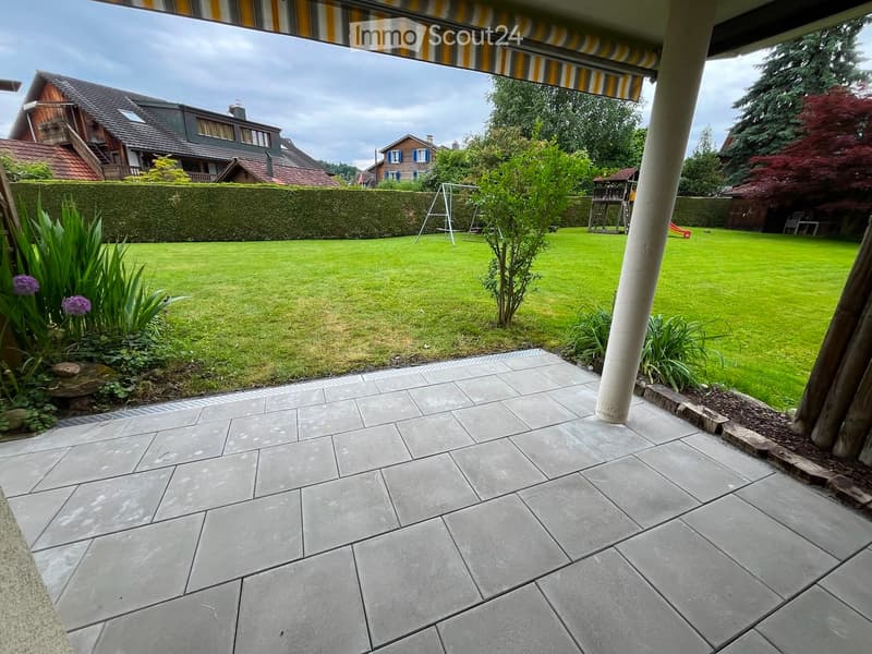 Well-maintained garden with a tiled patio, green lawn, hedge border, and surrounding trees and houses.