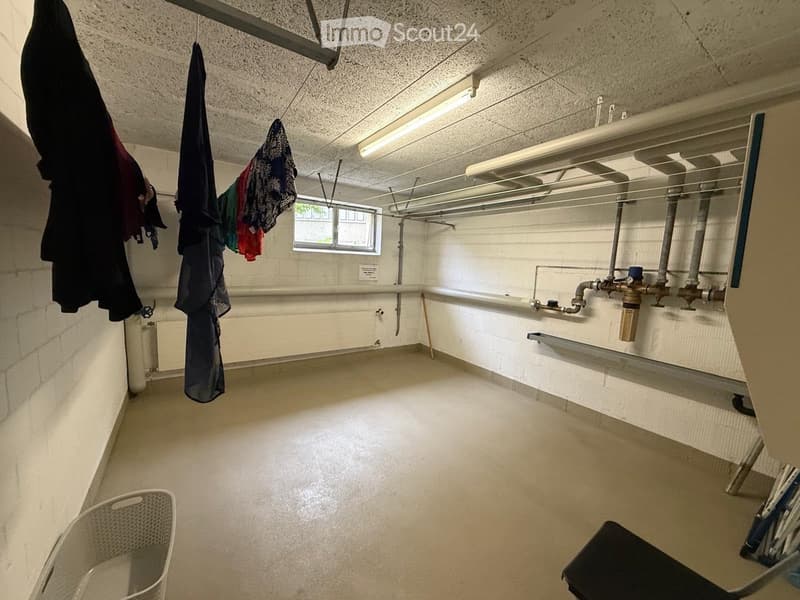Empty utility room with white tiled walls, concrete floor, exposed pipes, and a small window. Clothes are hanging on a rack, and a laundry basket is visible.