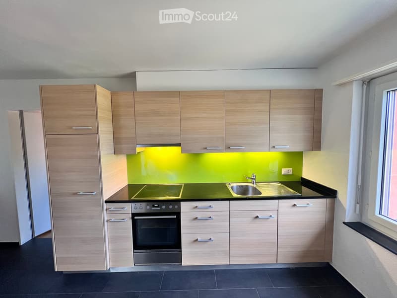 Modern kitchen with light wood cabinets, black countertops, and a green backsplash. Features an induction cooktop, oven, and sink.