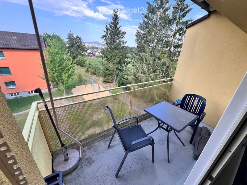 Balcony with a small table and two chairs, overlooking a green area with trees and a playground.