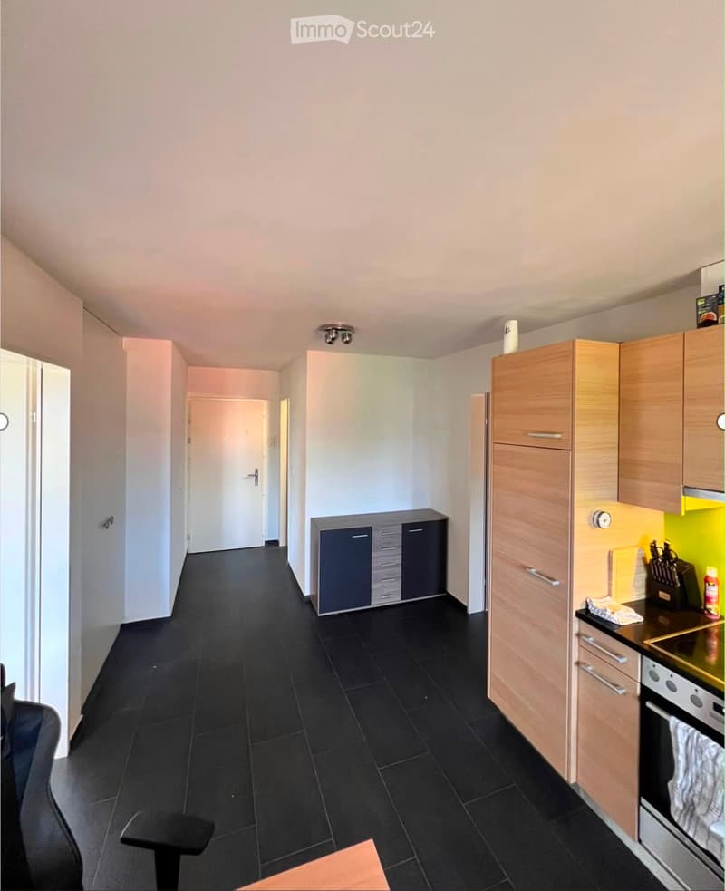 Modern kitchen with light wooden cabinets, black tiled floor, and stainless steel appliances including an oven. A small table and chair are visible in the foreground.