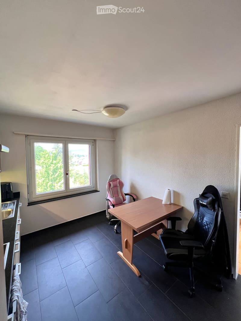 A small, furnished room with a wooden desk, two chairs, and a window offering a view of greenery outside.