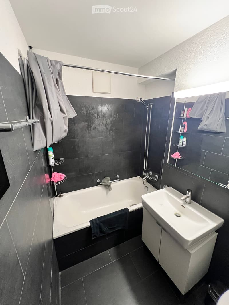 Modern bathroom with a bathtub, sink, mirror, towel rack, and shower. Dark tiles on walls and floor, with towels and toiletries visible.