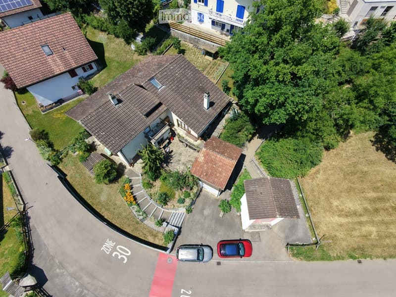 Aerial view of a residential area with multiple houses, gardens, and parked cars. The main house has a tiled roof, a balcony, and a driveway with two cars. Surrounding houses have similar architectural styles and well-maintained gardens.