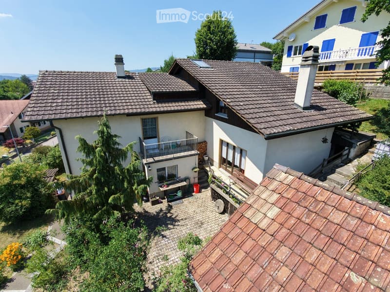 Two-story house with a tiled roof, multiple chimneys, and a balcony. The house is surrounded by greenery and has a small garden area with a table and chairs.