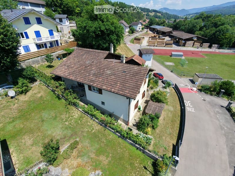 Aerial view of a residential area with multiple houses, green lawns, and a sports field. The houses have varied architectural styles, including pitched roofs and balconies. There are parked cars and a small playground visible.