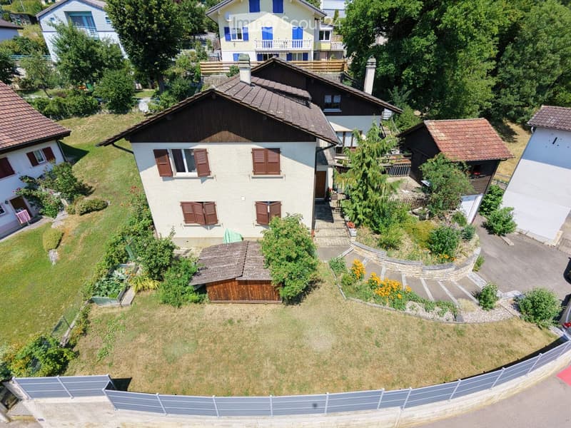 Aerial view of a two-story house with a beige facade, brown shutters, and a tiled roof. The house is surrounded by a well-maintained garden with various plants and trees. A wooden shed is located near the house, and the property is enclosed by a fence.