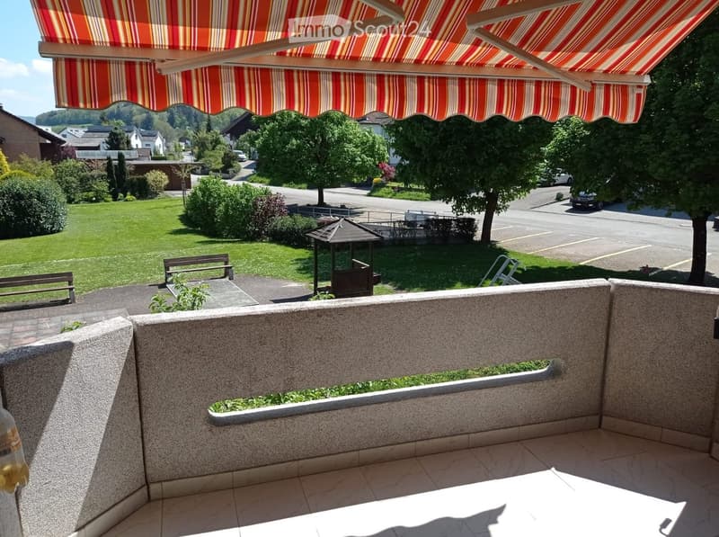 Balcony with tiled floor, stone railing, and striped awning, overlooking a green lawn and residential area.