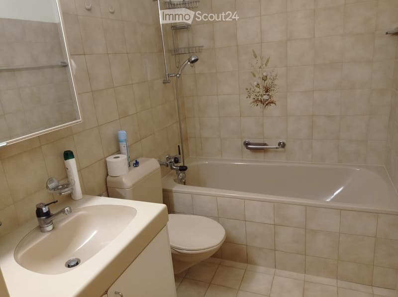 Bathroom with beige tiles, a sink, toilet, bathtub, and a showerhead. Toilet paper and a bottle are placed on the sink counter.