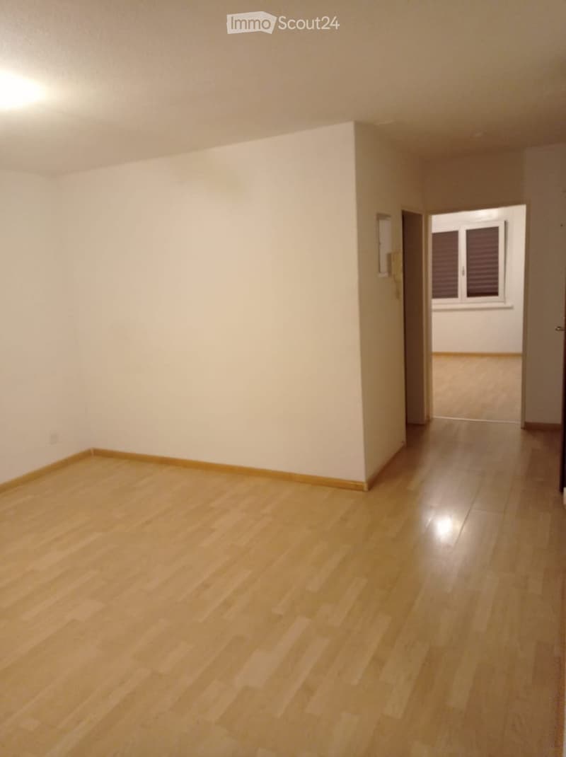 Empty room with light wooden flooring and white walls, featuring a doorway leading to another room with a window and tiled floor.