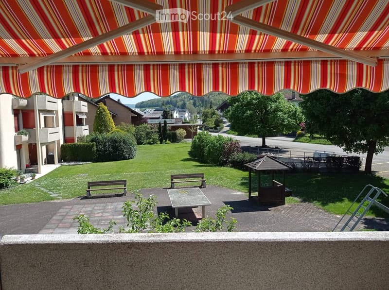 Balcony with striped awning, outdoor table, benches, and a small gazebo overlooking a green lawn and residential area.