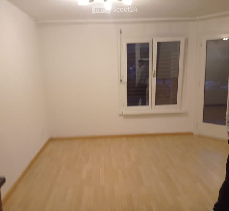 Empty room with light wooden flooring and white walls, featuring large windows with blinds and a small shelf below one window.