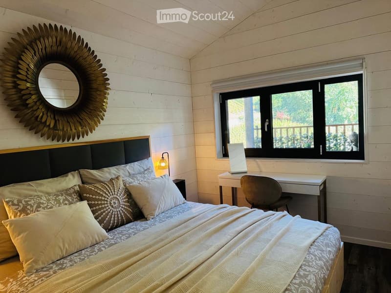 Cozy bedroom with a wooden bed, patterned pillows, a large round mirror with a sun-shaped frame, a small desk with a chair, and a window with a view of greenery.