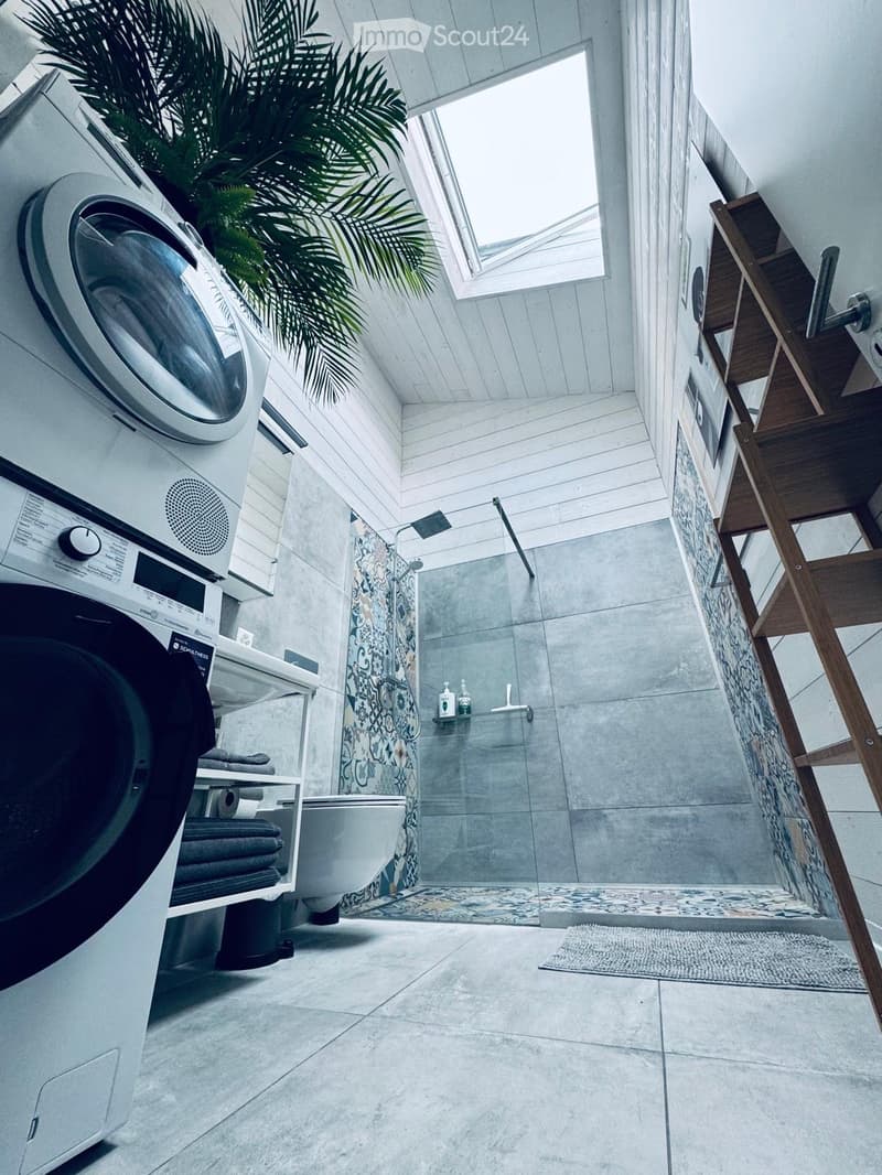 Modern bathroom with a washing machine, toilet, bathtub, and shower area. Features a wooden shelf, a potted plant, and a patterned shower curtain.