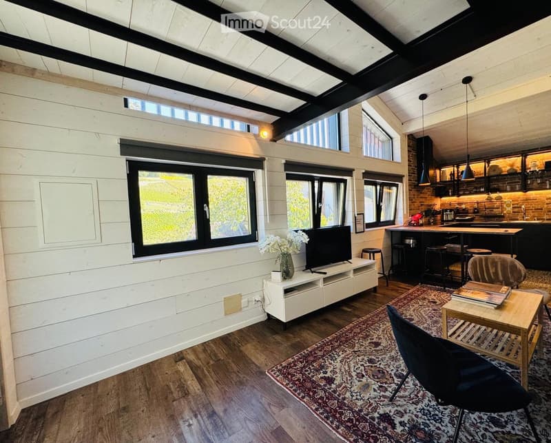 Open-concept living and dining area with exposed wooden beams, large windows, and a modern kitchen in the background.