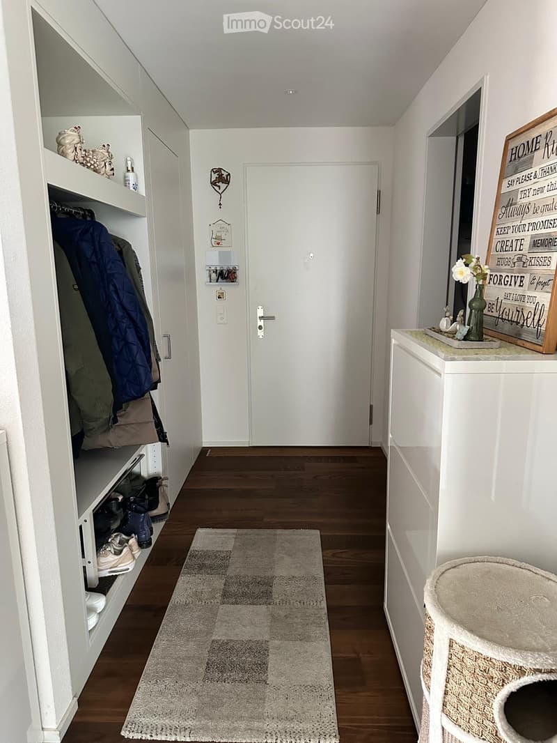 A well-lit hallway with wooden flooring, white walls, built-in storage with shoes and jackets, a small rug, and a decorative sign on a side table.