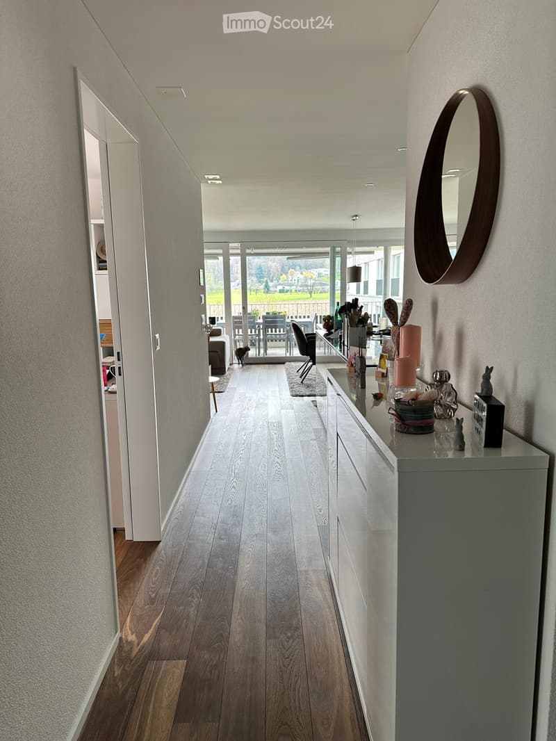 Modern hallway with light wooden flooring, white walls, and a small console table with decorative items. A large round mirror is mounted on the wall, and a sliding glass door leads to an outdoor area with furniture and a view of greenery.