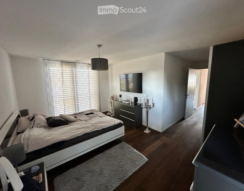 Modern bedroom with a large bed, nightstand, chair, small table, television mounted on the wall, candle holder, and a soft rug on the wooden floor.
