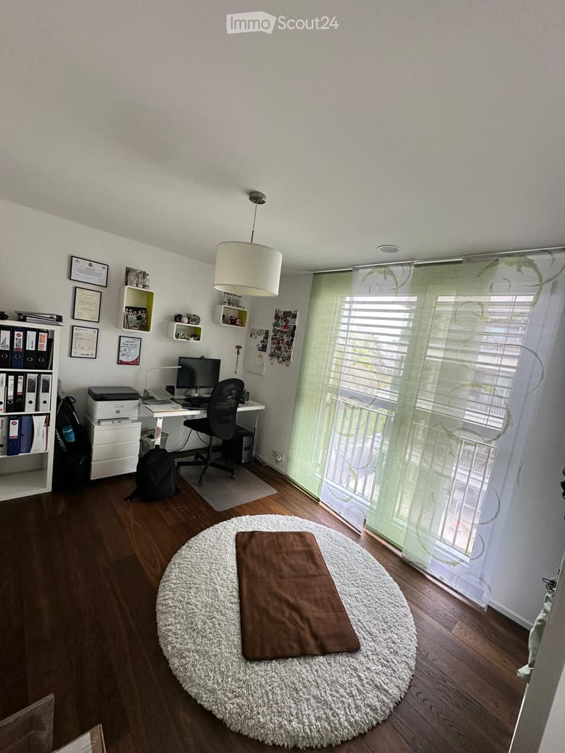 Well-lit room with wooden flooring, a round white shag rug, a brown cushion, a desk with a computer, a chair, a bookshelf, and a hanging lamp.