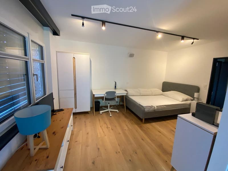 Cozy bedroom with a double bed, desk, chair, wardrobe, and nightstand. Features wooden flooring and large windows with blinds.