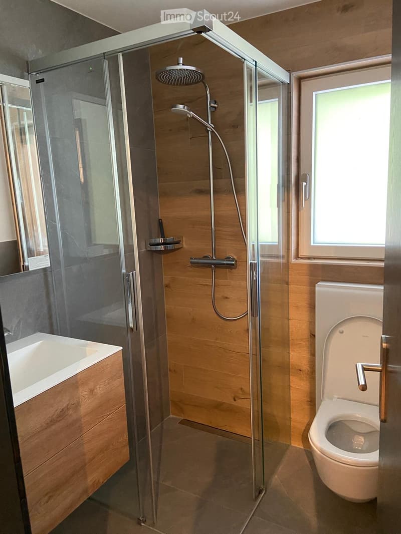 Modern bathroom with a glass-enclosed shower, a bathtub, and a toilet. The shower features a handheld showerhead and a fixed overhead showerhead. The walls are finished in wood and grey tiles.