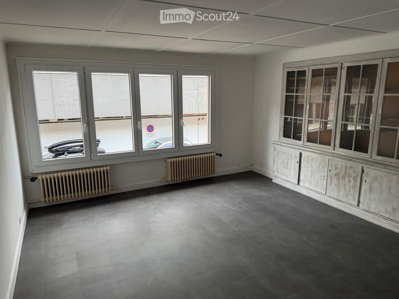 Empty room with grey tiled floor, white walls, large windows with blinds, radiator, and built-in cabinet with glass doors.