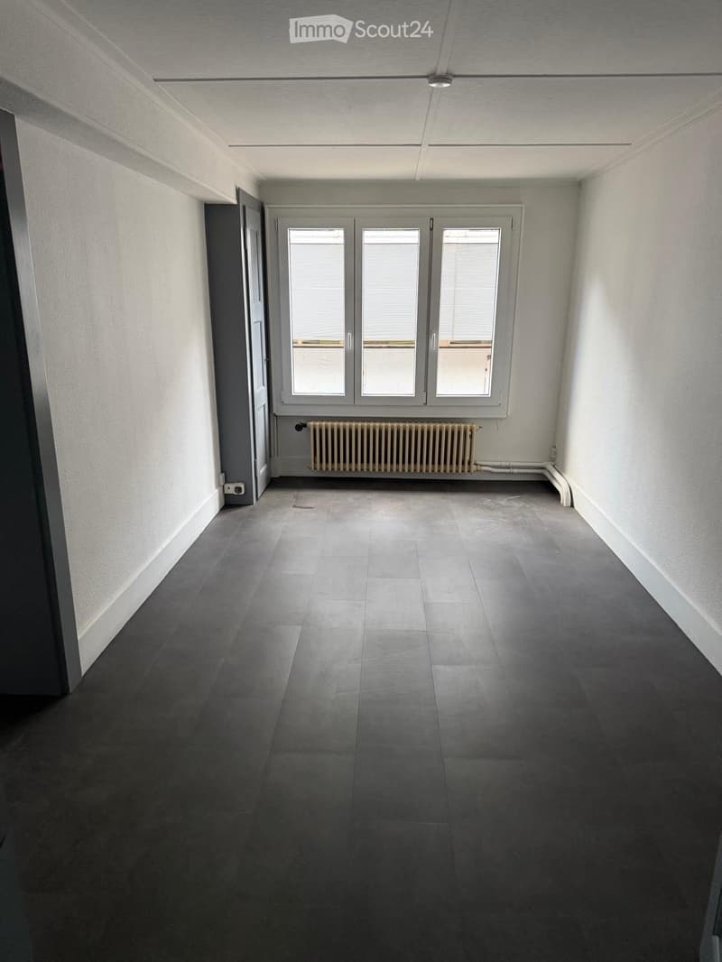 Empty room with grey tiled floor, white walls, large windows with blinds, radiator, and a door leading to another space.