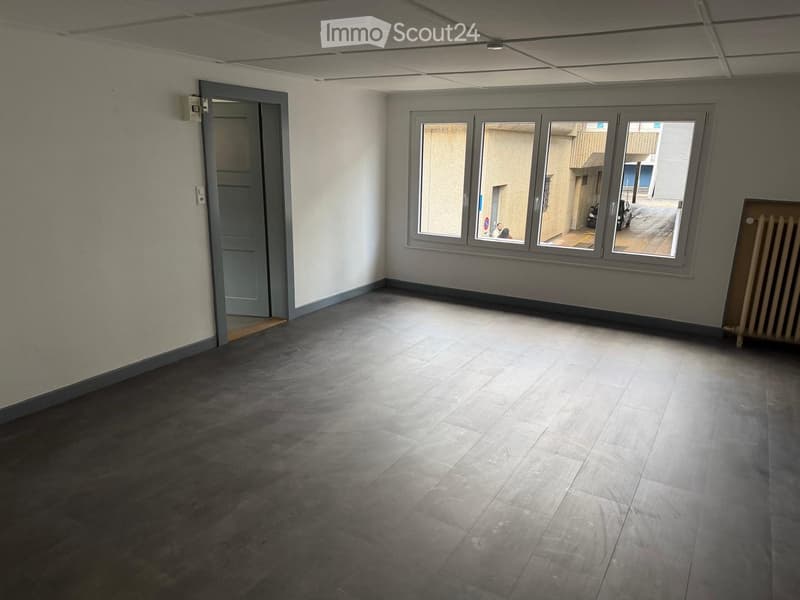 Empty room with grey tiled floor, white walls, grey door, large window with grey frame, radiator, and visible electrical outlet.