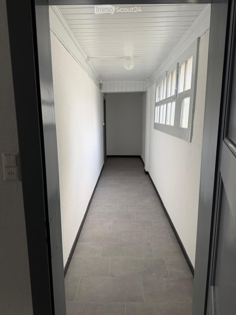 Empty hallway with tiled floor, white walls, and a single light fixture on the ceiling. Windows on the right side allow natural light to enter.