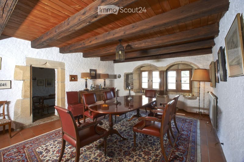 Elegant dining room with exposed wooden beams, stone walls, and large windows. Features a round wooden dining table surrounded by red leather chairs, lamps, and artwork.