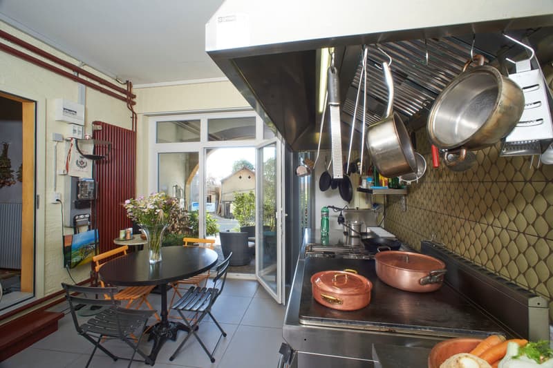The image shows a modern kitchen with stainless steel appliances, including a stove, oven, and refrigerator. Copper pots and pans are placed on the counter. A dining area with a round table and chairs is visible, along with a vase of flowers. The kitchen