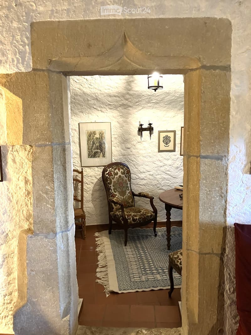 A small, cozy room with stone walls and a tiled floor, featuring a patterned armchair, a small wooden table, a wooden chair, and framed artwork on the walls.
