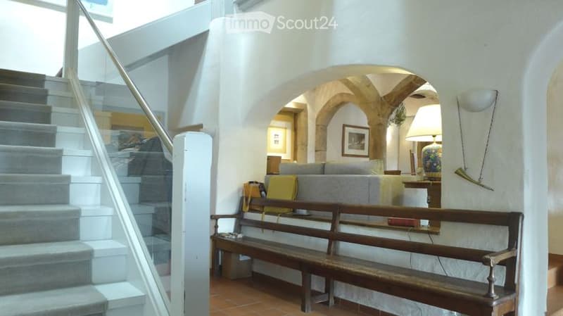 A modern hallway with a staircase on the left and an arched opening to a living area on the right. The living area features a grey sofa, a wooden bench, and a decorative lamp.