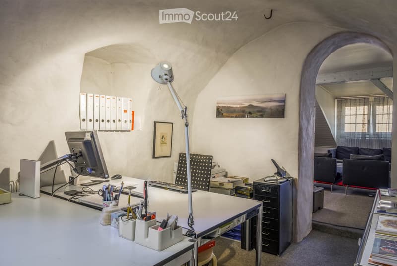 Office space with a large desk, computer setup, filing cabinets, and a small sitting area with a sofa. The room features exposed stone walls and arched doorways.