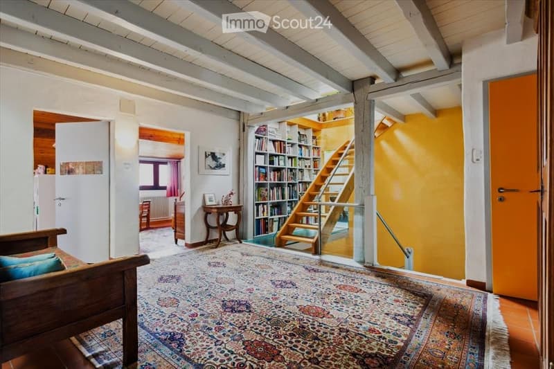 Bright living room with exposed wooden beams, colorful carpet, wooden sofa, small table, bookshelf, and staircase leading to another level.