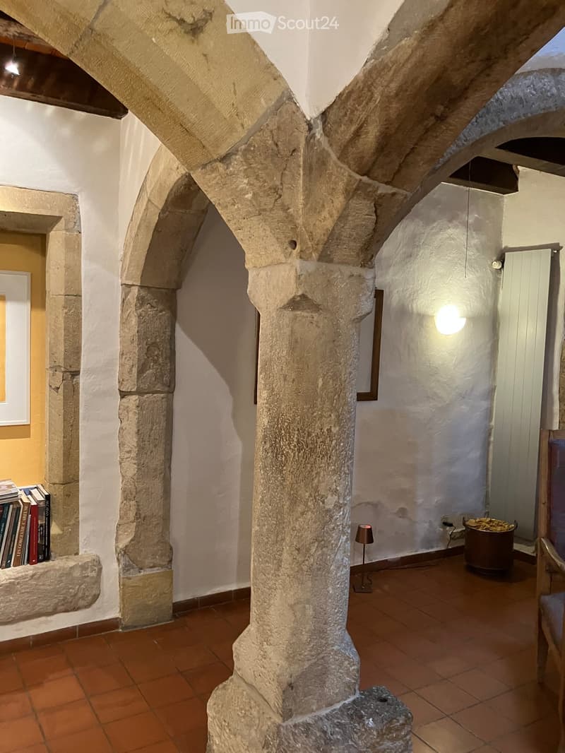 A rustic corridor with stone columns, terracotta tiles, and a small table lamp on the floor. Books are placed on a stone ledge, and a chair is visible in the background.