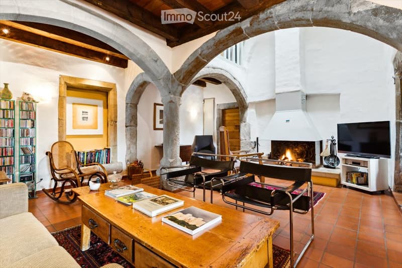 Living room with exposed wooden beams, terracotta tiles, fireplace, sofa, coffee table, rocking chair, bookshelf, and television.