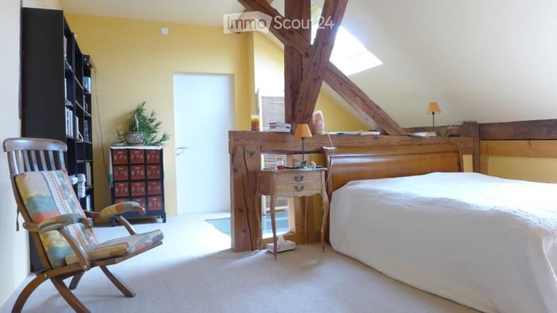 Cozy bedroom with wooden beams, a large bed with white bedding, a nightstand with a lamp, a wooden chair, a small plant, and a cabinet with drawers.