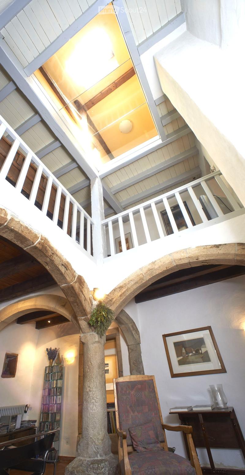 A spacious, well-lit room with exposed wooden beams and a stone column. Features include a patterned armchair, a small table with books and a glass vase, a bookshelf, and a decorative picture frame on the wall.