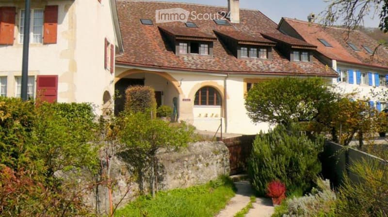 A charming two-story house with a red-tiled roof, white walls, and blue shutters. The house features arched windows and a small garden with neatly trimmed hedges and various plants.