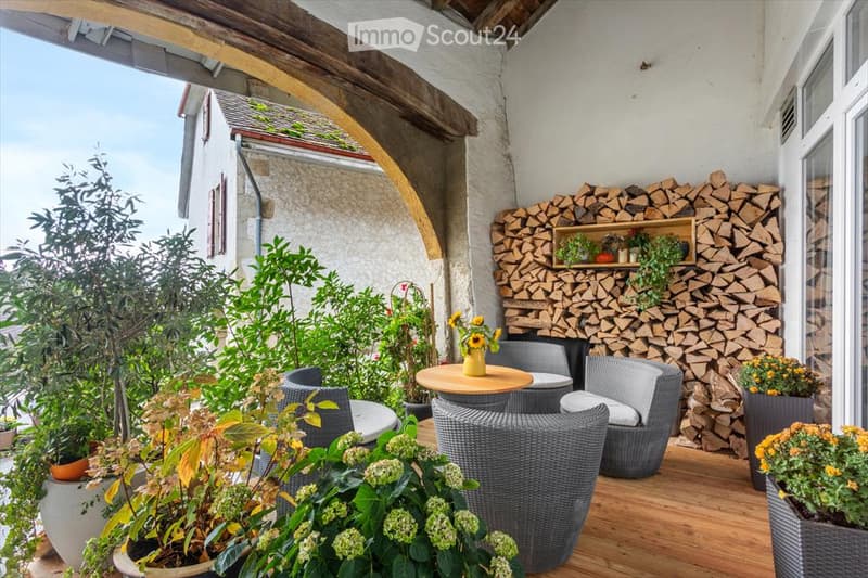 Wooden outdoor patio with wicker chairs, round table, potted plants, and a woodpile fireplace.