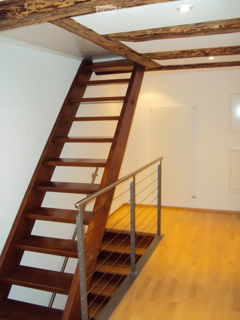 Wooden staircase with metal railing, exposed wooden beams on the ceiling, light-colored walls, and wooden floor.