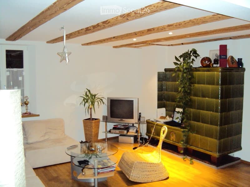 Living room with exposed wooden beams, fireplace, sofa, coffee table, wicker chair, ottoman, television, and plants.