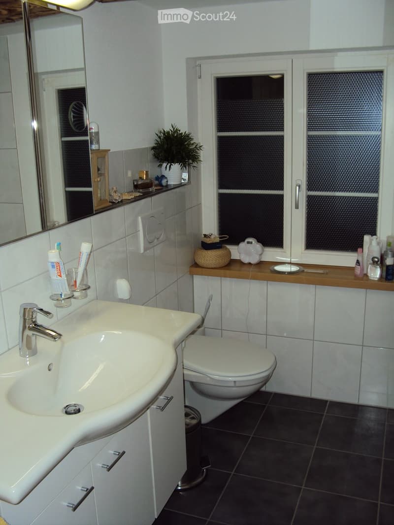 Modern bathroom with white tiles, a white sink with a chrome faucet, a toilet, a large mirror, and a window with frosted glass. The countertop holds a plant, a decorative bowl, a decorative plate, and various toiletries.